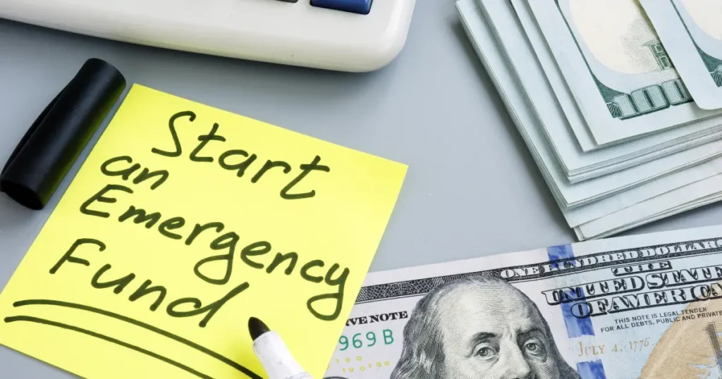 Yellow sticky note reading "Start an Emergency Fund" next to a stack of US hundred dollar bills and a calculator