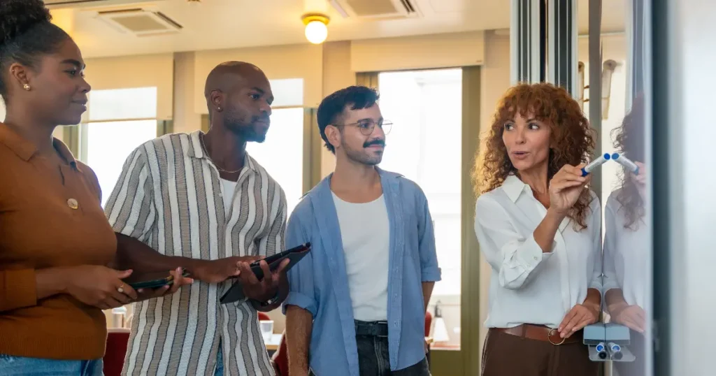 Woman writing on a whiteboard explaining rules to a diverse group of three people in a casual indoor setting