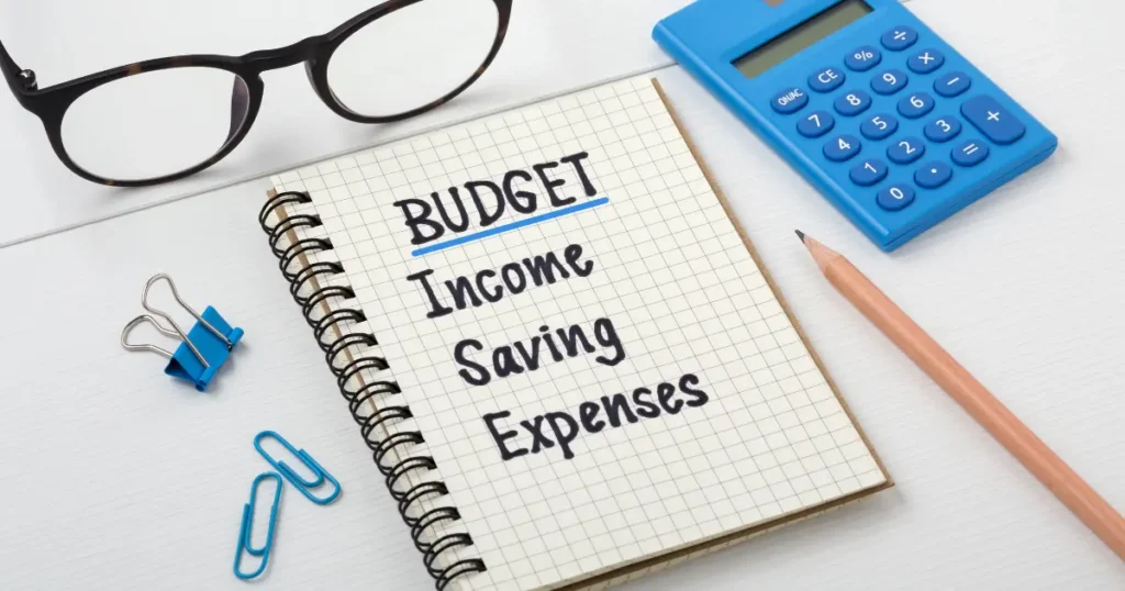 Notebook with budget, income, saving, and expenses written out alongside a calculator, pencil, and glasses — financial planning basics