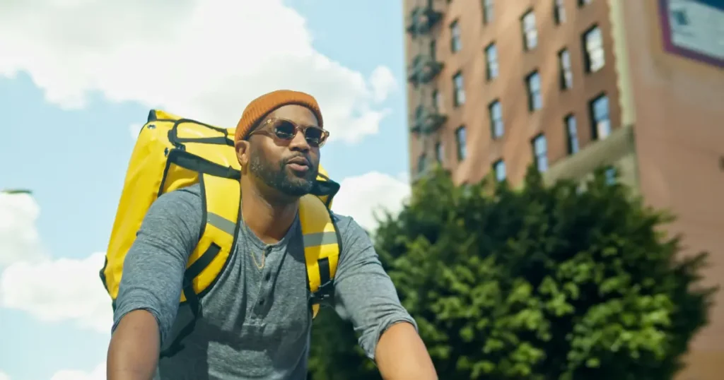 Black man on a bicycle with a large yellow delivery backpack in an urban city setting