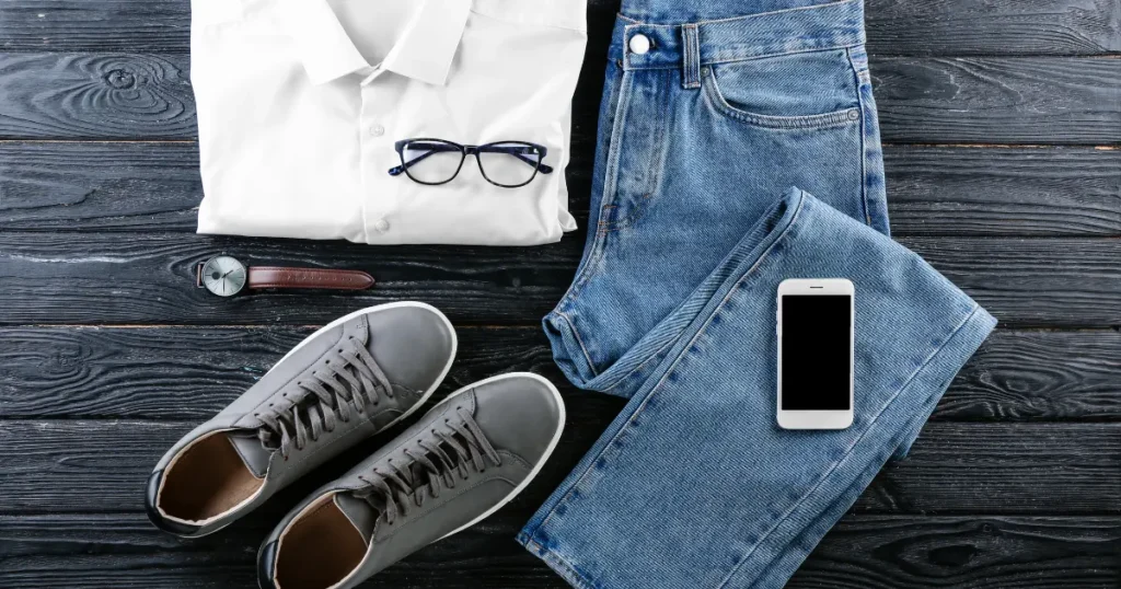 Flat lay of job-ready clothing — white button shirt, jeans, gray sneakers, watch, and glasses on a dark wood surface