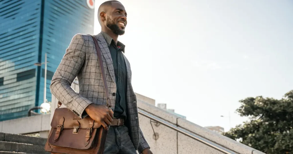 Well-dressed Black man in a blazer carrying a leather bag walking up steps outside an urban building