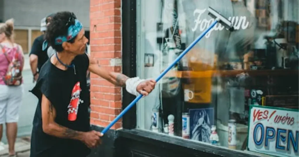 Person cleaning the front window of a small storefront with an Open sign visible inside