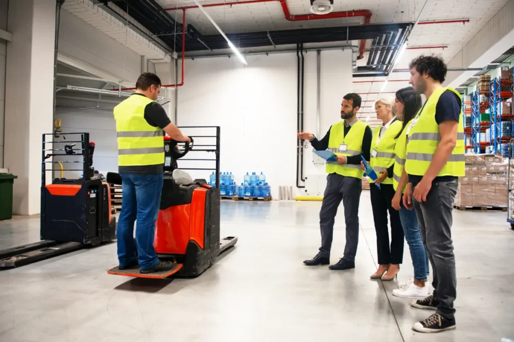 Workers in yellow safety vests observing a forklift operator being trained in a warehouse setting