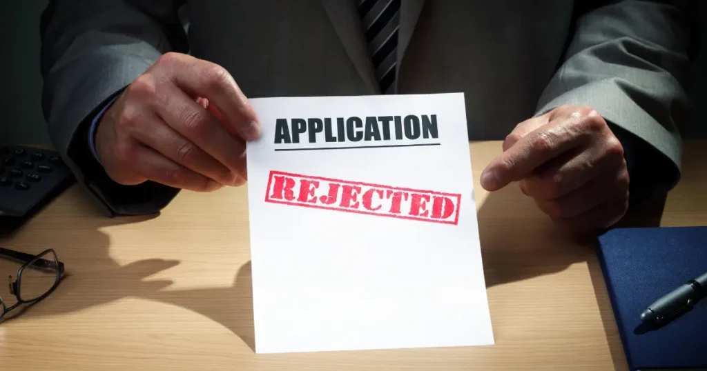 Person in a suit holding a document stamped with "Application Rejected" across a desk