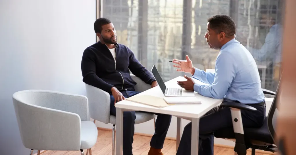 Two Black men in a serious conversation across a desk with a laptop in a professional office setting