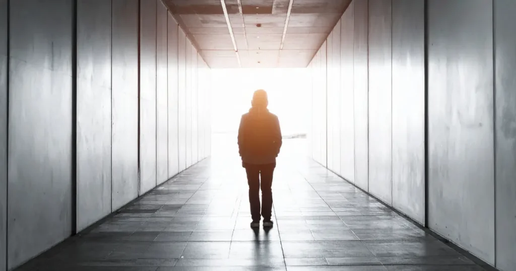 Silhouette of a person standing alone in a concrete corridor facing bright light at the end