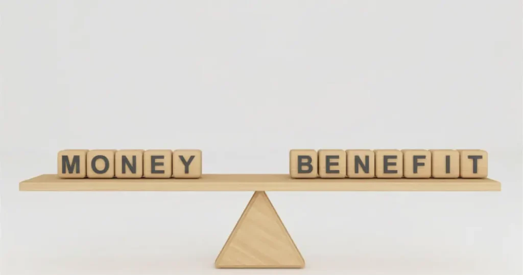 Wooden letter blocks spelling "Money" and "Benefit" balanced on opposite ends of a seesaw scale