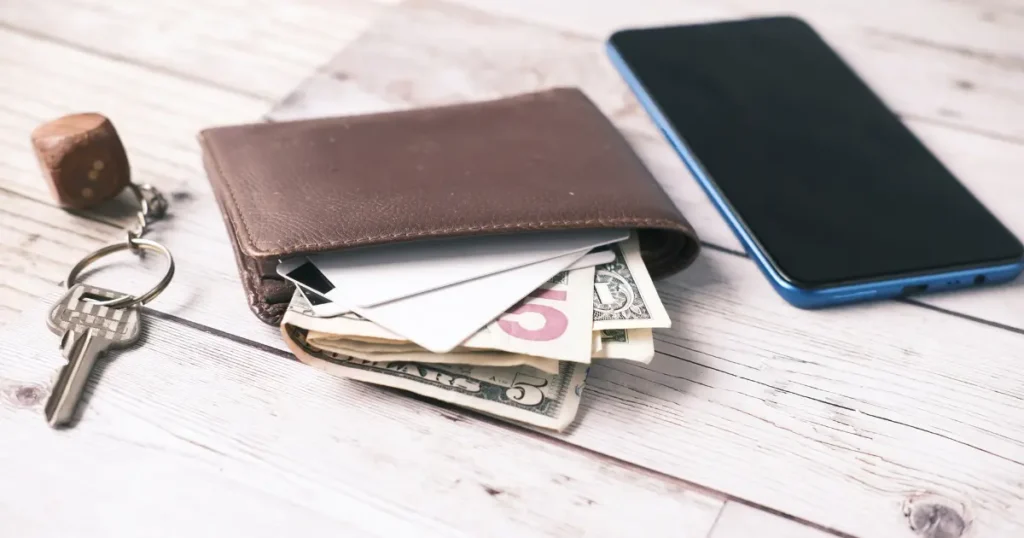 Leather wallet with cash and cards, house keys, and a smartphone on a wooden surface