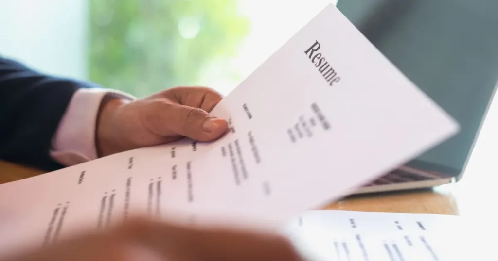 Person in a suit holding a resume document with a laptop visible in the background