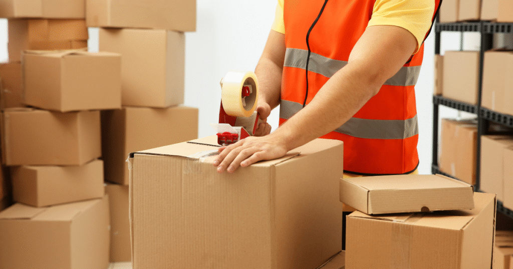Warehouse worker in an orange safety vest taping cardboard boxes surrounded by stacked packages